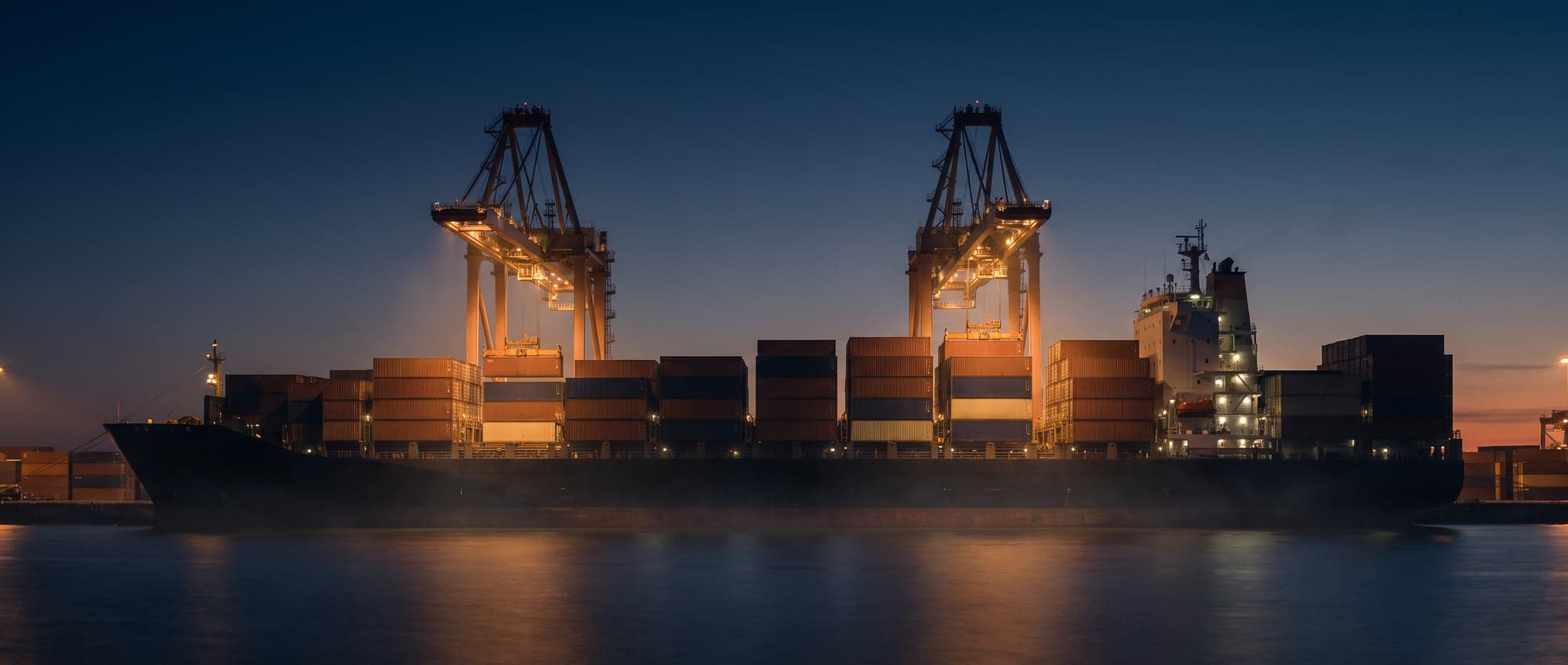 Containerterminal an einem Fluss in der Abenddämmerung mit beleuchteten Kränen